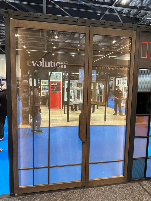A large glass display door with dark bronze metal framing and a vertical handle, showing a showroom interior with blue flooring and people reflected or visible through the glass. The word 'evolution' is printed on the glass.