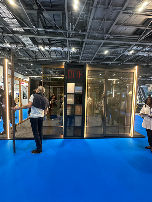A glass and metal modular display booth or cabin with a black central frame and red 'OTIF' lettering, shown at an exhibition. Several people are standing around it, and the floor is bright blue.